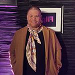 Javi Simons stands in front of a purple wall. He wears a black t-shirt, jeans, a brown overcoat, and a colorful scarf. 他在微笑.