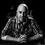 Grad Terry Watson sits with his hands clasped at a desk. He is wearing a plaid button-down shirt and is in a music studio.